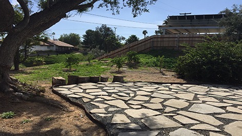 Stone pathway leads through a landscaped yard with a large tree, grassy areas, and a wooden fence in the background. The setting is calm and inviting.